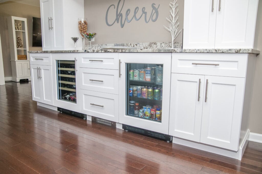 A home bar area with white cabinets, a granite countertop, and two glass-front mini fridges stocked with beverages. The word Cheers is displayed on the wall above the counter.