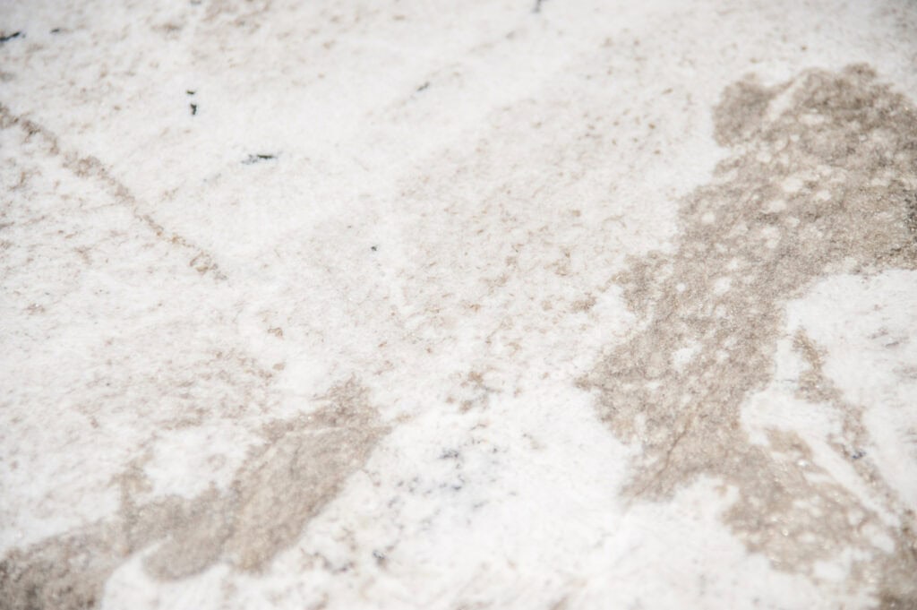 White marble surface with subtle light brown and gray veining, creating a natural and textured pattern. The surface appears smooth with slight color variations and organic lines.