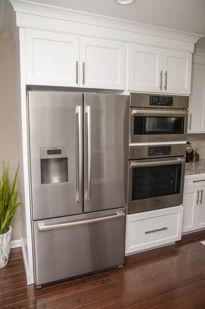 A modern kitchen features a stainless steel French door refrigerator with a water dispenser, built-in microwave, oven, and white cabinets, all set on dark wood flooring.