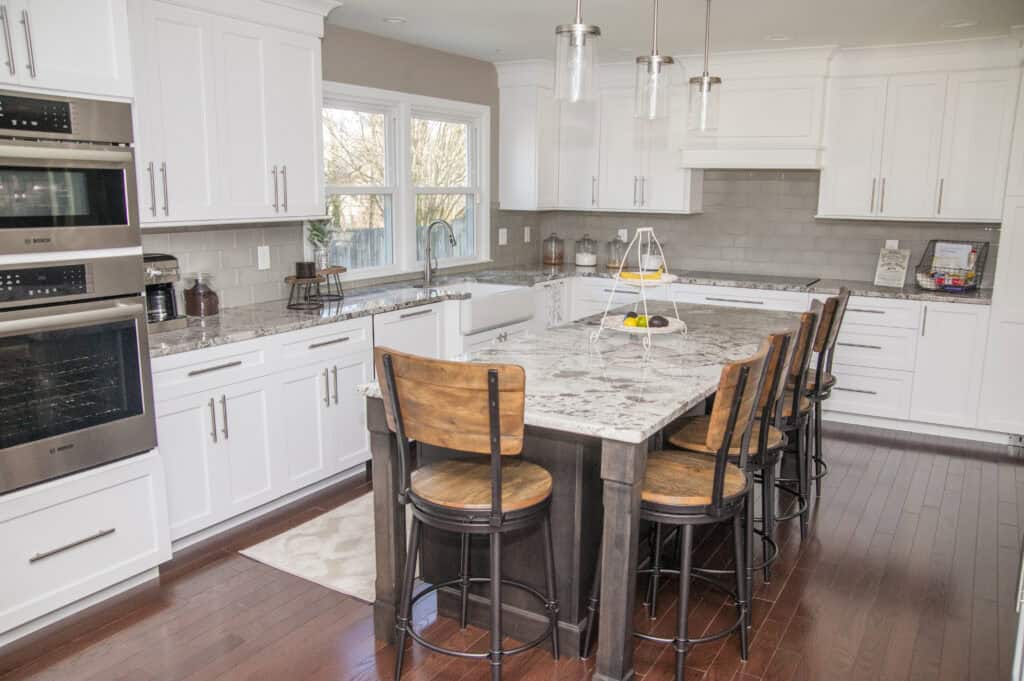 Modern kitchen with white cabinets, granite countertops, stainless steel appliances, and a large island with four wooden bar stools on dark hardwood floors. Pendant lights hang above the island. Sunlight streams through the window.