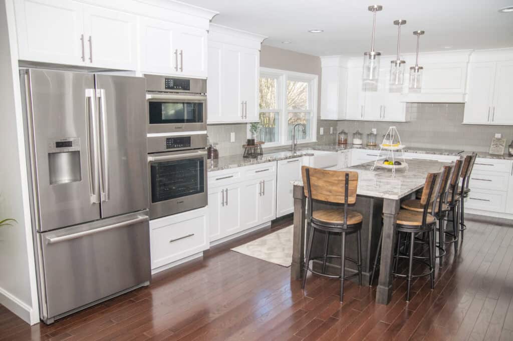 Modern kitchen with white cabinets, stainless steel appliances, a large island with a marble countertop, four wooden bar stools, hardwood floors, and large windows letting in natural light.