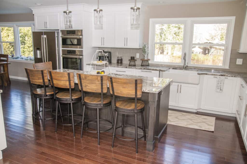 Bright, modern kitchen with white cabinets, stainless steel appliances, granite countertops, and a large island with five wooden barstools. Hardwood floors and large windows provide natural light.