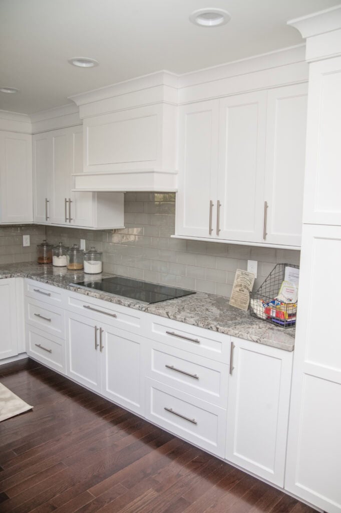 A modern kitchen with white cabinets, stainless steel handles, gray stone countertops, glass jar canisters, a metal basket with snacks, and a glossy gray subway tile backsplash on dark wood flooring.