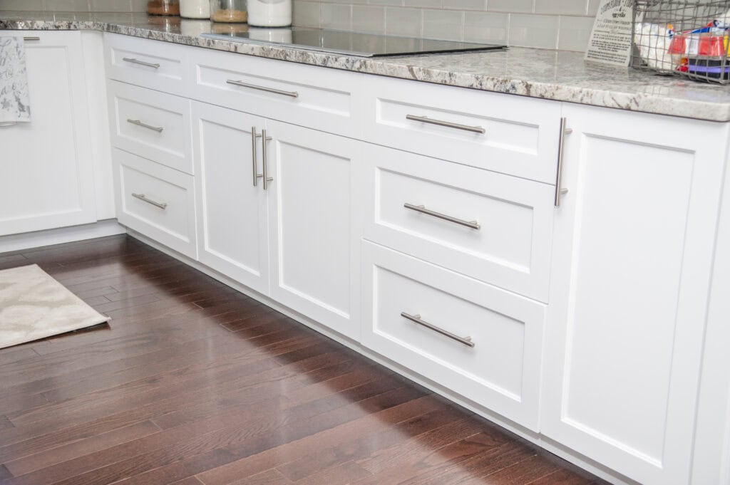 White kitchen cabinets with silver handles are installed below a marbled countertop. The floor is dark wood, and household items are visible on the counter, including containers and a wire basket.