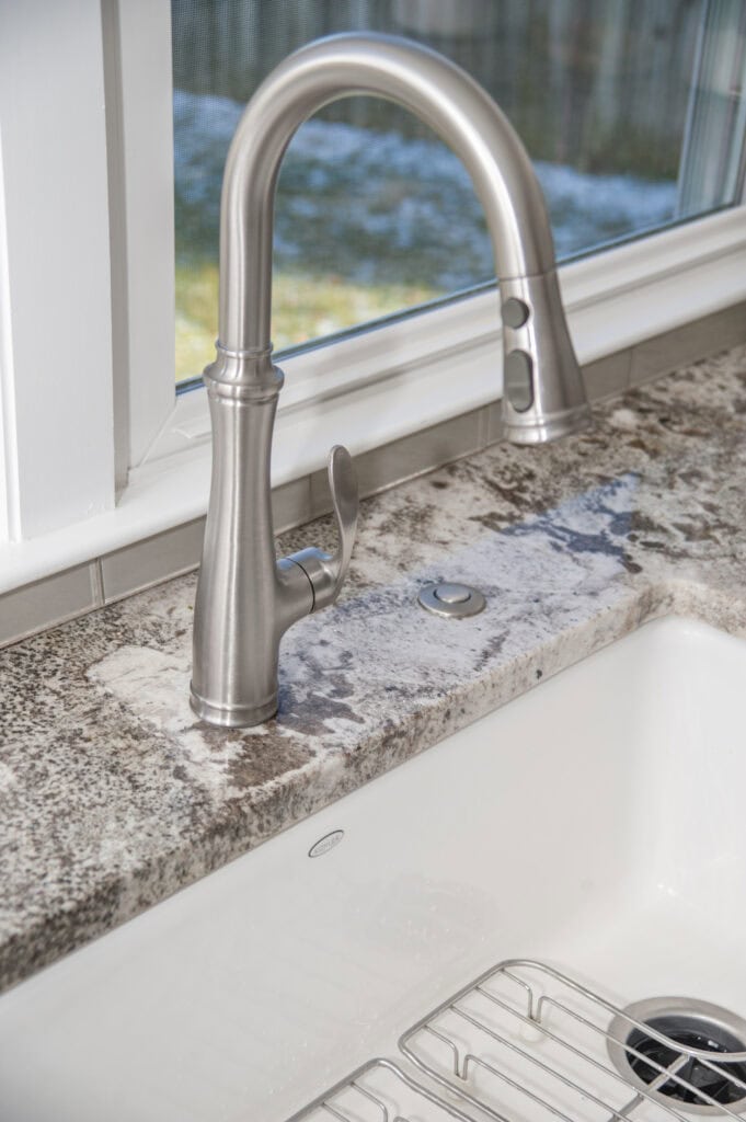 A close-up of a modern stainless steel kitchen faucet with a curved spout, set in a granite countertop, beside a white sink and a window showing an outdoor view.