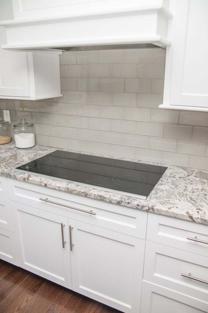 Modern kitchen with white cabinets, a speckled granite countertop, a sleek black electric cooktop, and a beige subway tile backsplash. Two glass jars with dry goods sit on the counter to the left.