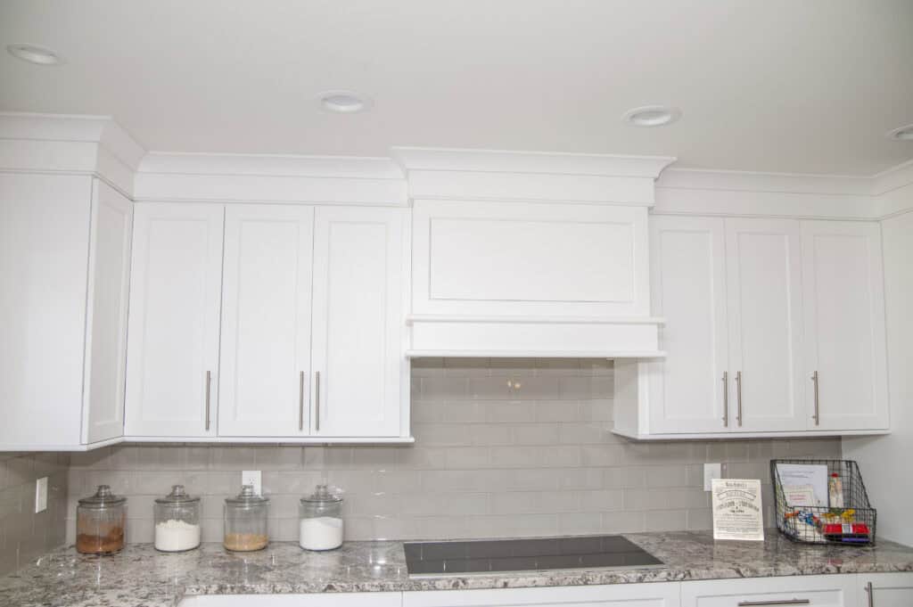 A modern kitchen with white cabinets, a light gray tiled backsplash, and a granite countertop. Three jars with pantry items sit on the counter, along with a framed certificate and a rack of colorful condiment packets.