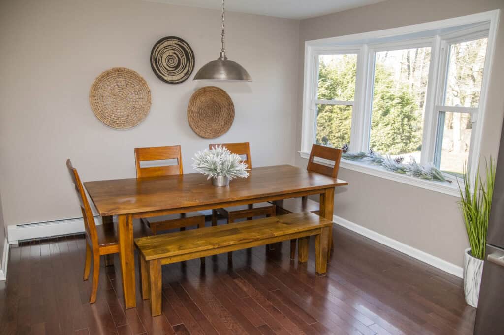 A dining room with a wooden table, three wooden chairs, and a matching bench. The table has a white plant centerpiece. Three woven wall decorations hang above, and a large window lets in natural light. A tall plant is near the window.