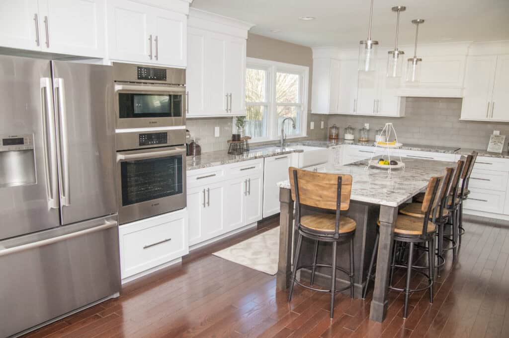 Modern kitchen with white cabinets, stainless steel appliances, double ovens, a large island with a marble countertop, four wooden bar chairs, pendant lights, and hardwood floors. Sunlight streams in through two windows.