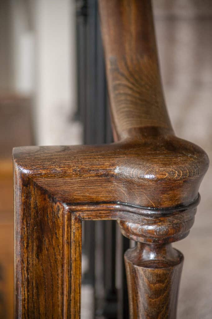 Close-up of a polished wooden handrail and banister post on a staircase, showing rich wood grain and smooth, glossy finish with blurred background.