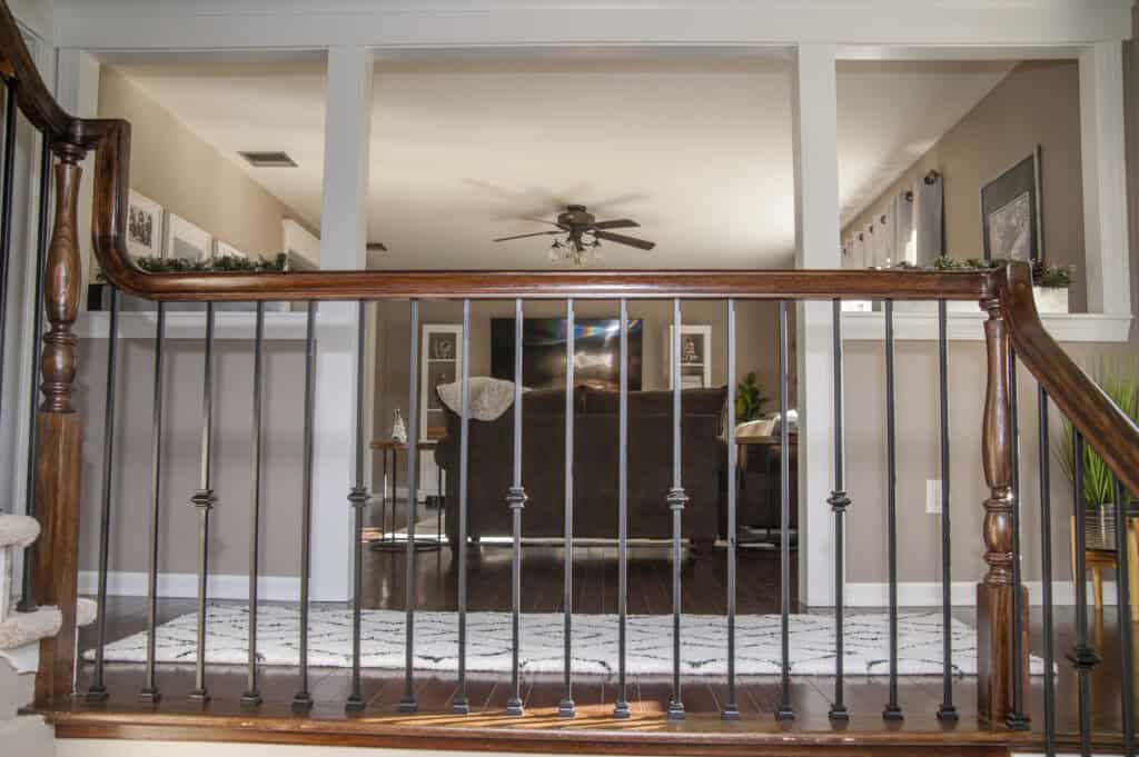 View of a living room from behind a wooden and black metal stair railing, featuring a brown couch, TV, ceiling fan, framed pictures, and a striped rug on dark wood floors.