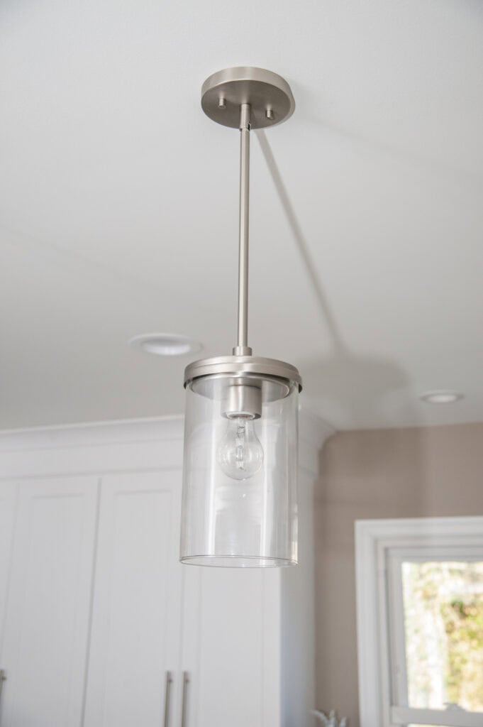 A modern ceiling pendant light with a cylindrical glass shade and an exposed bulb, mounted on a metal rod, hangs from a white ceiling in a bright room with white cabinetry and a window.