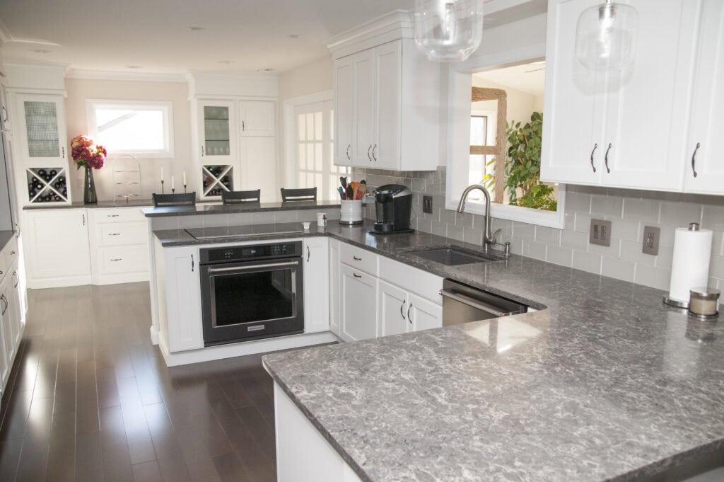 Modern kitchen with white cabinets, gray marble countertops, stainless steel appliances, a built-in oven, a coffee maker, and a window overlooking a bright, plant-filled room.
