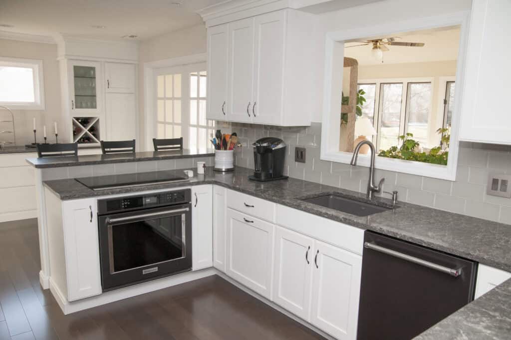 Modern kitchen with white cabinets, dark countertops, a built-in oven, a sink, a coffee maker, and utensils. A window above the sink looks into a bright sunroom with plants and large windows.