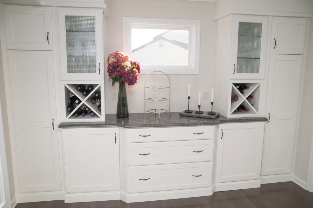 A white built-in cabinet with wine storage, glass-fronted upper shelves, gray countertop, two candles, a tiered stand, and a vase with pink and red flowers, set below a window.