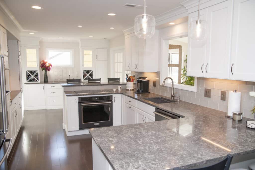Modern kitchen with white cabinets, gray marble countertops, an island with a built-in stove, stainless steel appliances, pendant lights, and bar stools at a breakfast bar. A vase with flowers sits in the background.