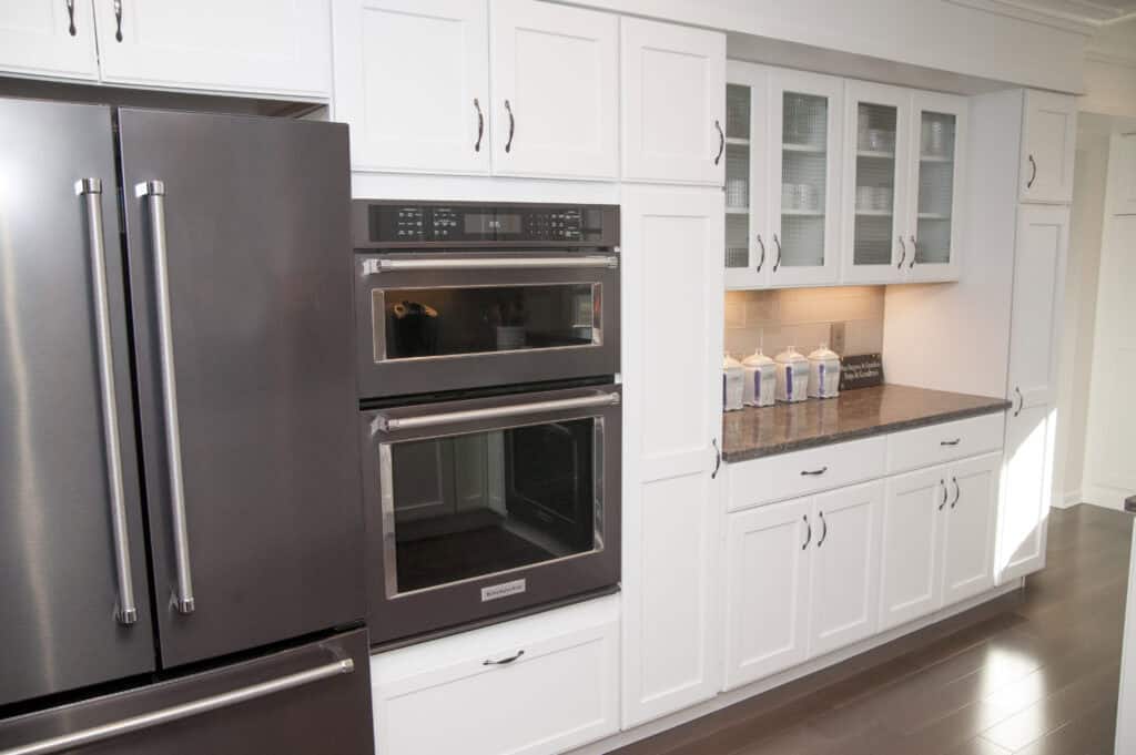 Modern kitchen with white cabinets, stainless steel double oven, and a large refrigerator. The counter has decorative jars, and there are glass-front upper cabinets above the counter.