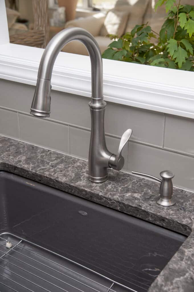 A modern stainless steel kitchen faucet and soap dispenser are installed on a dark granite countertop next to a deep black sink, with green plants and a bright room visible in the background.