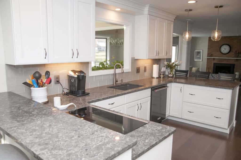 Modern kitchen with white cabinets, gray marble countertops, built-in stove, coffee maker, sink, and utensils in a holder. Pendant lights hang over the counter, and a window opens to a dining area in the background.