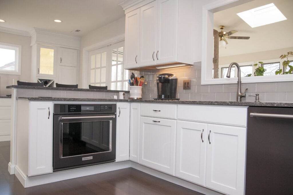 Modern kitchen with white cabinets, stainless steel oven, gray countertops, a sink, and a coffee maker. Bright natural light streams through windows and a large interior pass-through window.