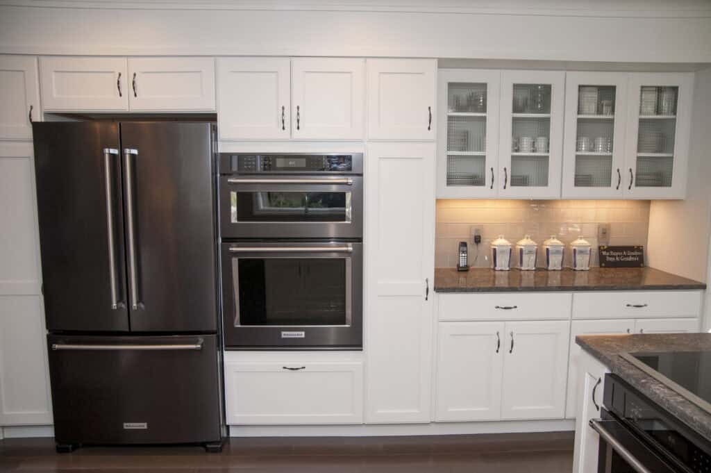 Modern kitchen with white cabinets, stainless steel refrigerator, double wall oven, and a countertop with glass jars and an electric outlet. Glass-front cabinets display neatly arranged dishes.