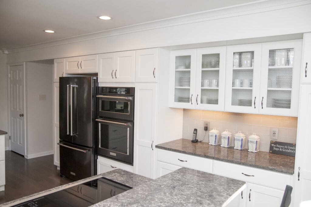 Modern white kitchen with dark countertops, stainless steel double oven, black refrigerator, glass-front cabinets, and a row of blue-and-white canisters on the counter. A landline phone is also visible on the counter.