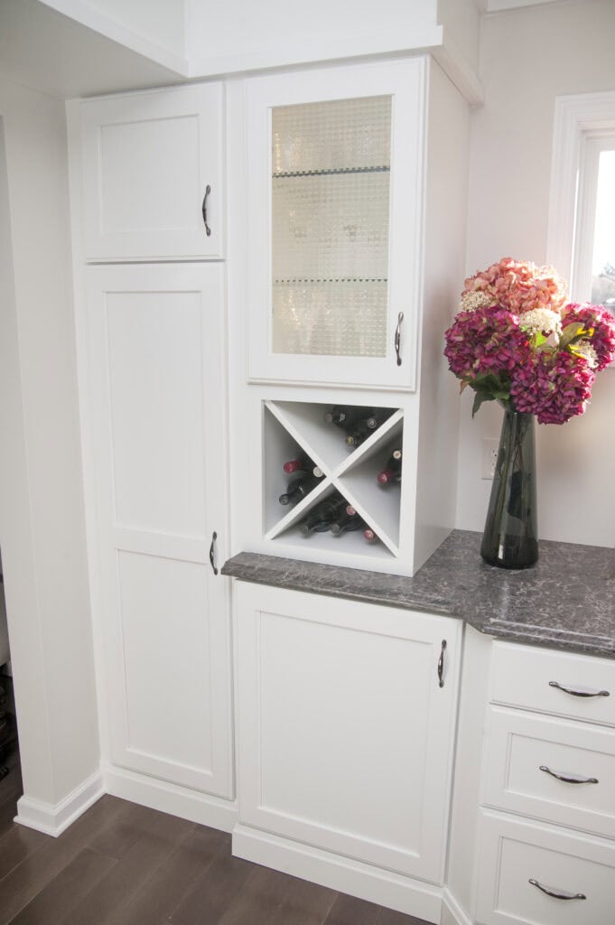 White kitchen cabinets with a gray countertop, a small wine rack holding bottles, glass-fronted upper cabinet, and a large vase of colorful hydrangeas placed on the counter near a window.