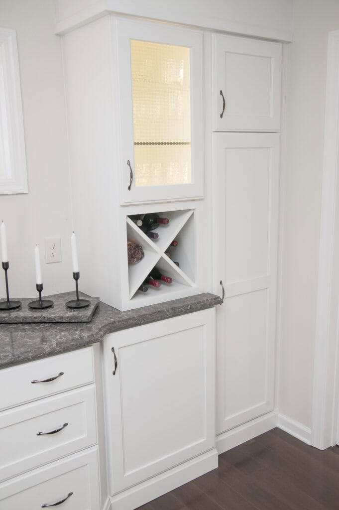 White kitchen cabinets with a gray countertop, a built-in wine rack holding several bottles, a glass-front upper cabinet with interior lighting, and a candle holder with three white candles on the counter.