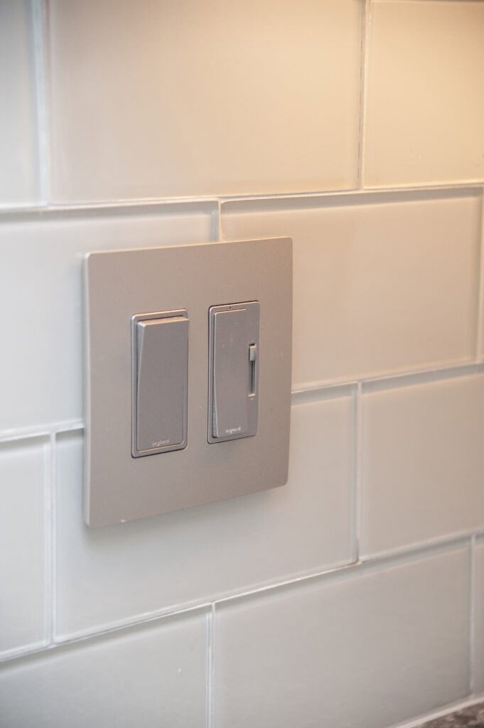 A close-up of a modern, gray light switch panel with two switches mounted on a white subway tile wall.