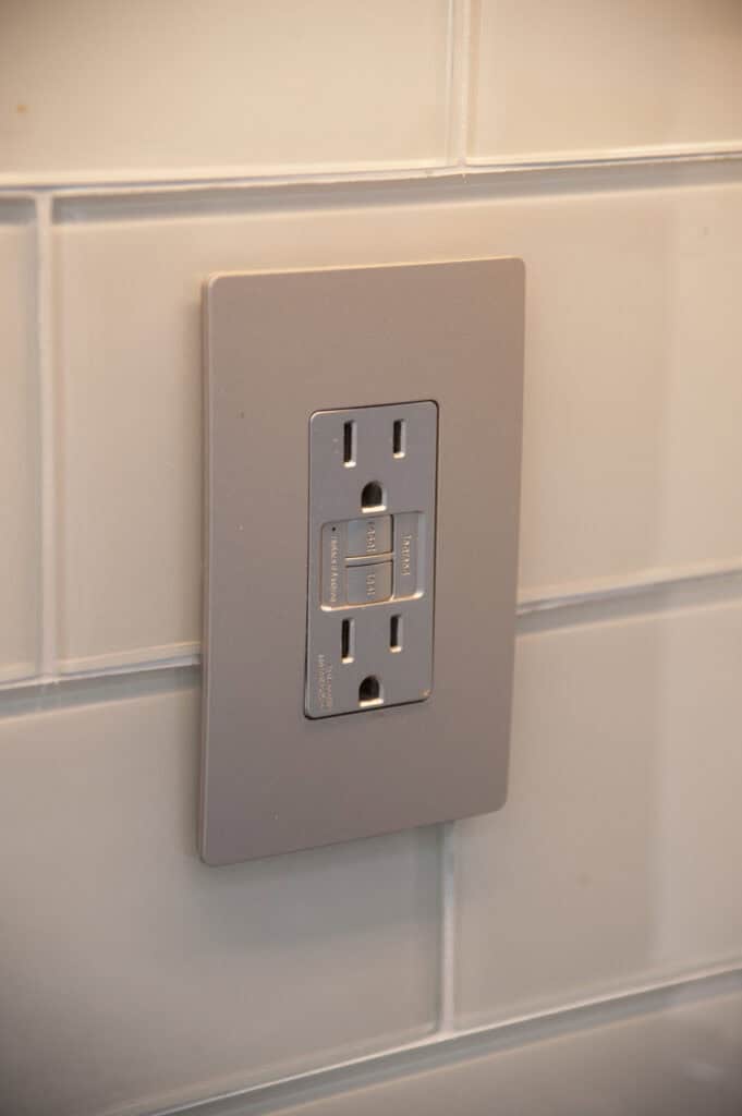 A close-up of a GFCI electrical outlet installed on a white tiled wall. The outlet has two sockets and reset/test buttons in the center.