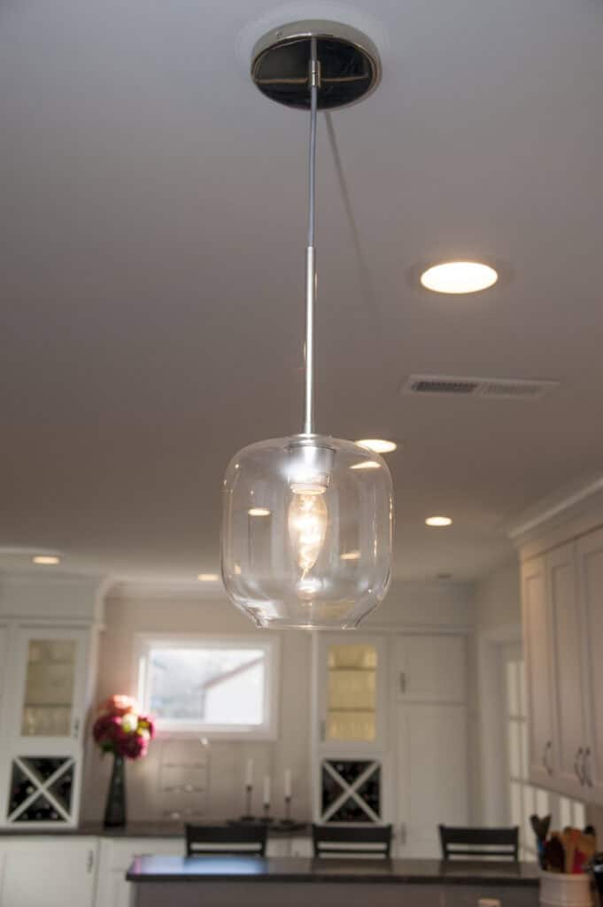 A modern pendant light with a clear glass shade hangs from the ceiling in a bright, contemporary kitchen with white cabinets, recessed lighting, and a vase of flowers on the counter in the background.