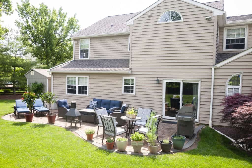 Backyard patio with outdoor furniture, potted plants, a grill, and a chiminea beside a beige house. There is a grassy lawn and lounge chairs, with trees and a shed in the background.
