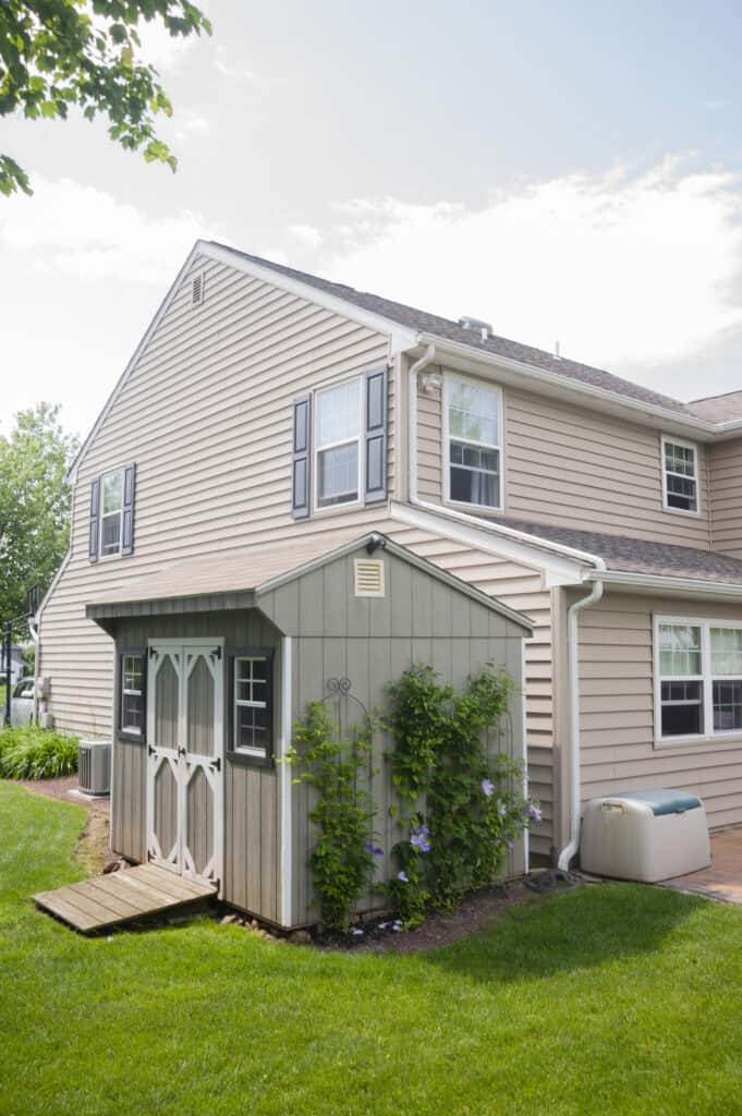 A beige two-story house with black shutters, a small gray shed attached, climbing plants on the shed, green grass, and a clear sky in the background.
