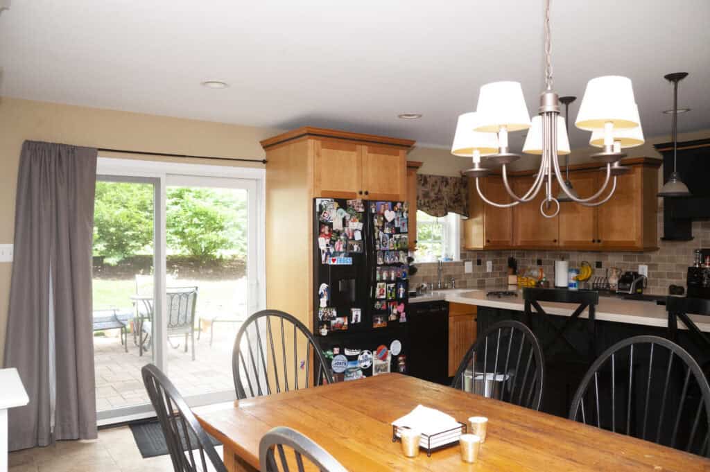 A bright kitchen with wooden cabinets, a black refrigerator covered in magnets, a wooden dining table with chairs, and a chandelier. Sliding glass doors lead to a patio with outdoor furniture and greenery visible outside.