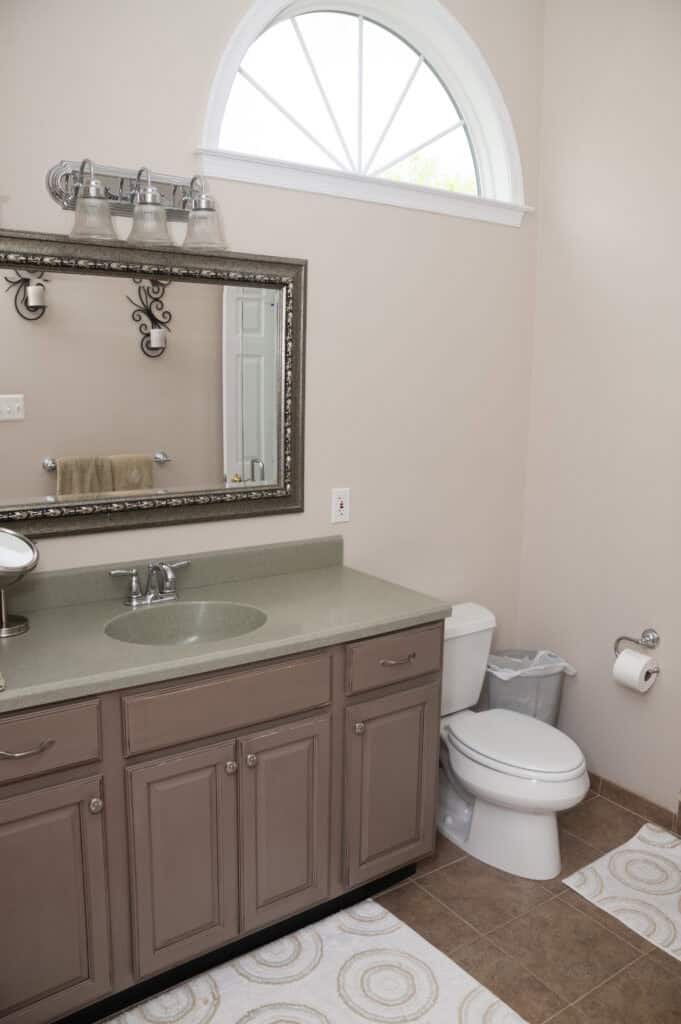 A bathroom with a sink, mirror, and brown vanity on the left, a toilet and trash bin on the right, a towel on a rack, and a decorative window above. The floor has tan tiles and patterned rugs.