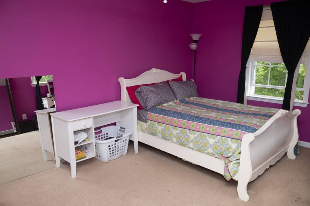 A bedroom with bright pink walls, a white bed with colorful bedding, a small white desk with shelves, a laundry basket, and windows with black and white curtains letting in natural light.