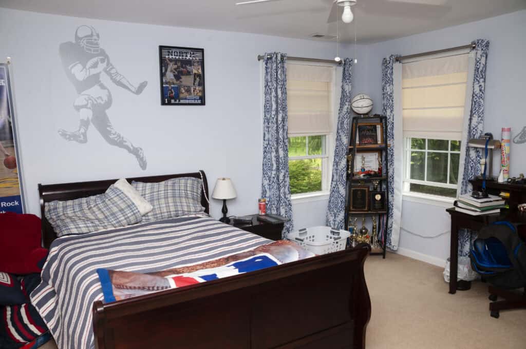 A neatly made bed with striped bedding in a sports-themed bedroom, decorated with a football player wall decal, trophies, books, a desk, and blue patterned curtains on two windows.