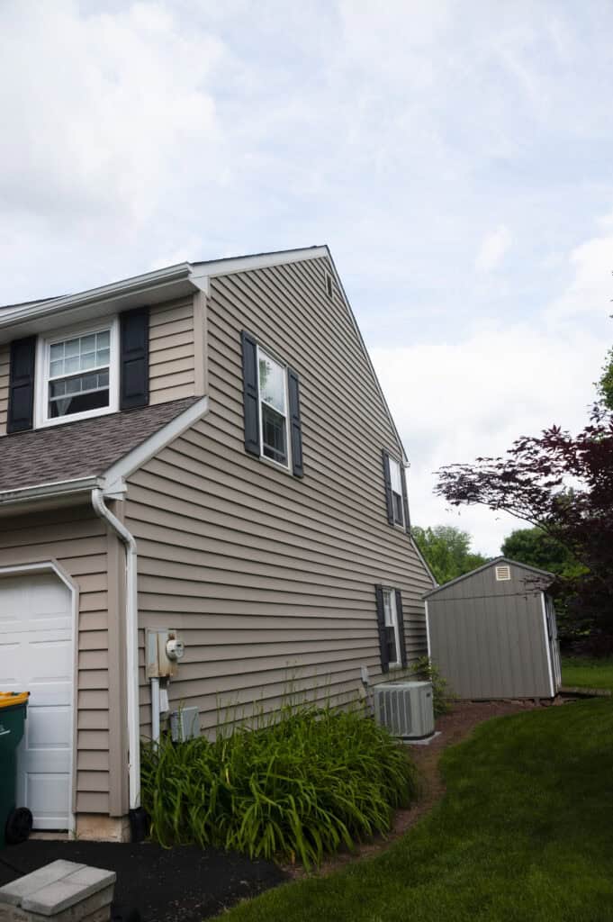 A beige-sided house with black shutters, white trim, and a sloped roof. There are green plants by the wall, an air conditioning unit, a small shed in the backyard, and some trees in the background under a partly cloudy sky.