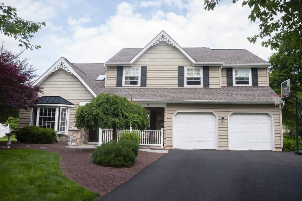 A two-story beige suburban house with white trim, a front porch, a double garage, and a well-kept yard with green grass, shrubs, and trees on a sunny day.