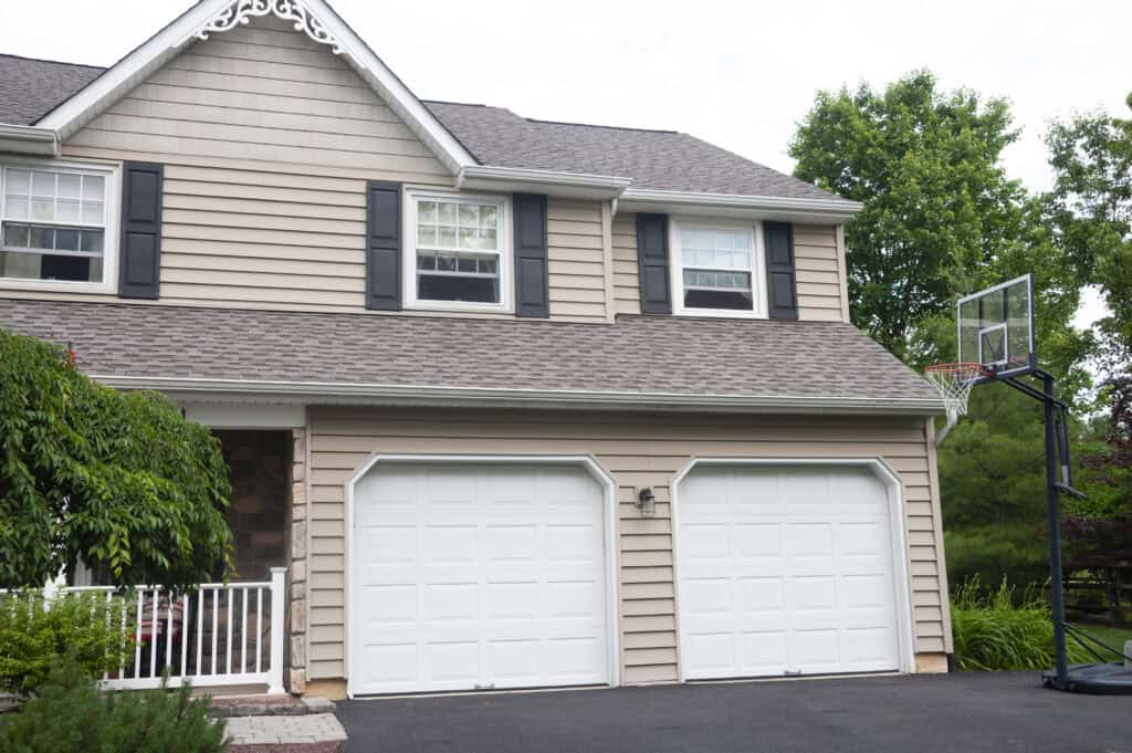 A suburban house with tan siding, black shutters, and two white garage doors. A portable basketball hoop stands on the driveway near green trees and bushes.