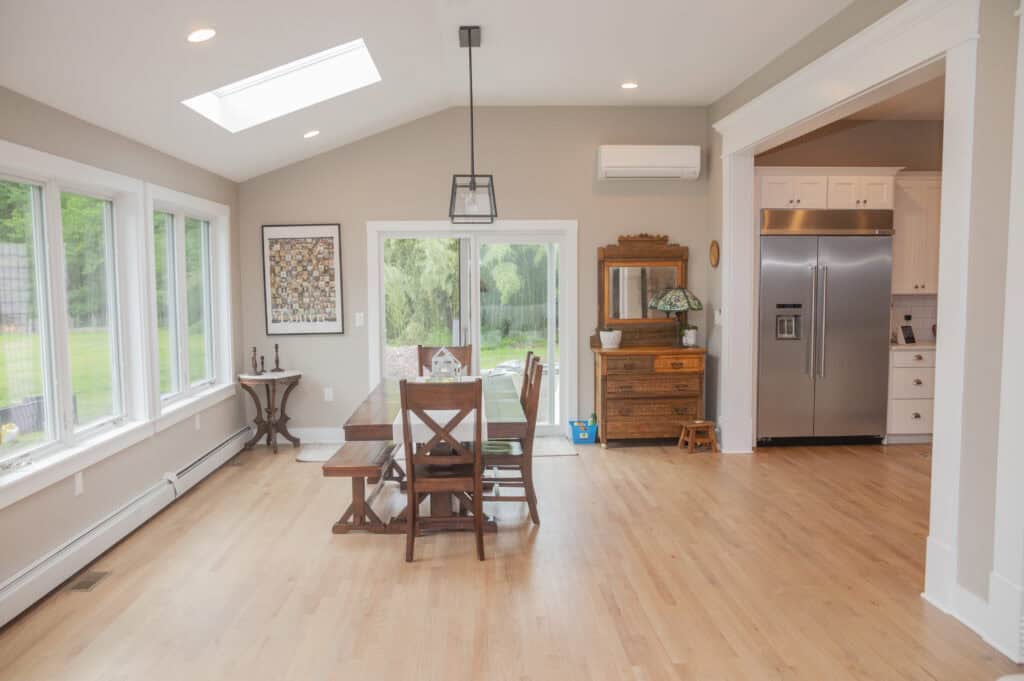 Bright dining room with wooden table and chairs, large windows, skylight, and sliding glass door; kitchen with stainless steel fridge visible through a wide doorway. Light wood floors and neutral walls throughout.