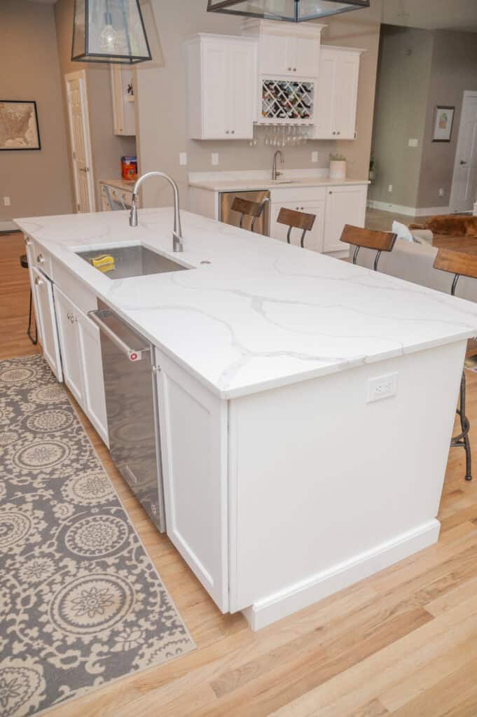 Bright modern kitchen with a large white marble island, built-in sink, dishwasher, and wooden bar stools. Light wood flooring and a gray patterned rug complete the space. White cabinets and wine storage are visible in the background.