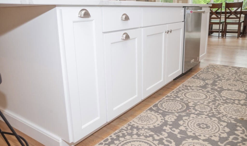 White kitchen cabinets with silver handles are shown under a white countertop. A stainless steel dishwasher is installed on the right. A patterned gray and white rug lies on the wooden floor in front of the cabinets.