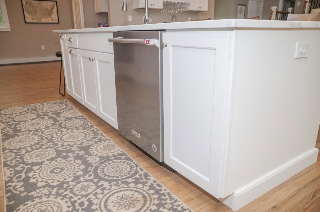 A stainless steel dishwasher is installed in a white kitchen island with panel detailing, next to a patterned gray and white rug on a wooden floor. The kitchen has light walls and visible decor in the background.