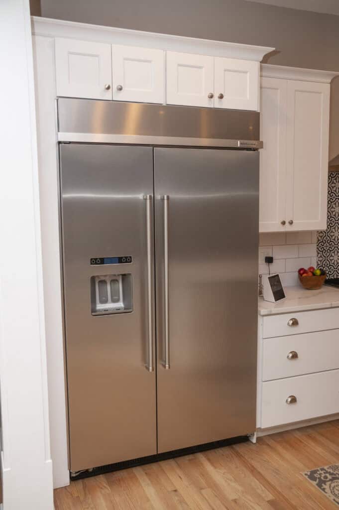 A modern stainless steel side-by-side refrigerator with double doors, an ice and water dispenser, and white cabinetry above and around it in a kitchen setting.