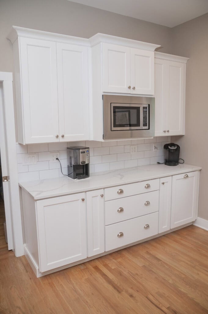 Modern kitchen with white cabinets, stainless steel microwave, coffee machine, and Keurig on a white countertop, white tile backsplash, and light wood flooring.