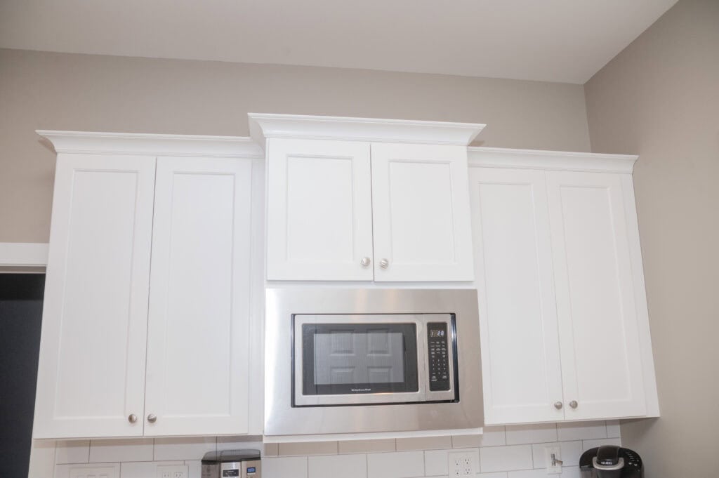White kitchen cabinets with silver knobs are mounted on a beige wall. A stainless steel microwave is built into the cabinetry above a white tile backsplash. A coffee maker and toaster are visible on the countertop below.