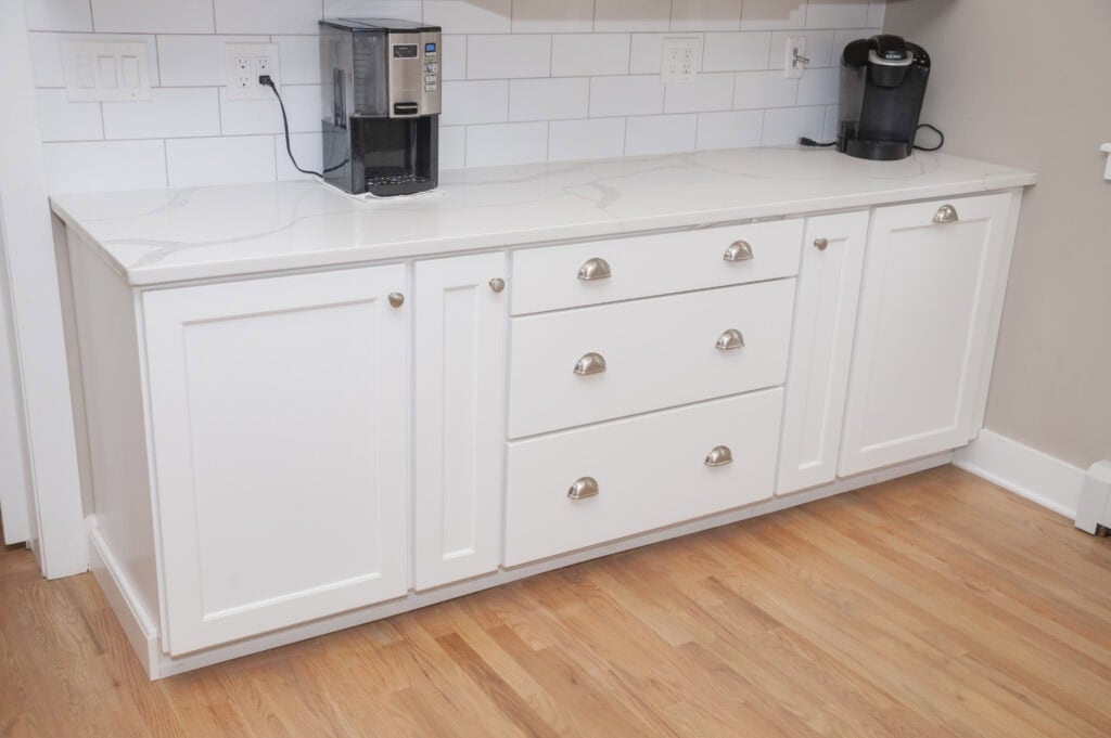 White kitchen cabinets with silver handles, three drawers in the center, and two doors on each side. The countertop is white with a coffee maker and a Keurig machine. The backsplash is white tile and the floor is light wood.