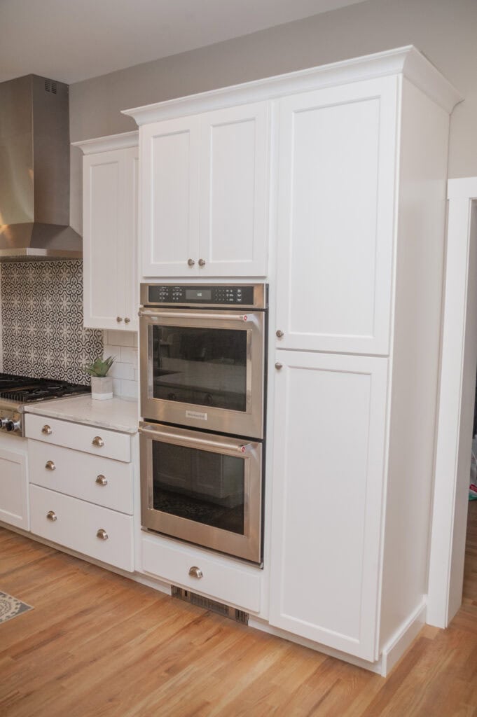 Tall white kitchen cabinets with silver handles, a built-in stainless steel double oven, and wood flooring. Part of a patterned backsplash and countertop with drawers are also visible on the left.