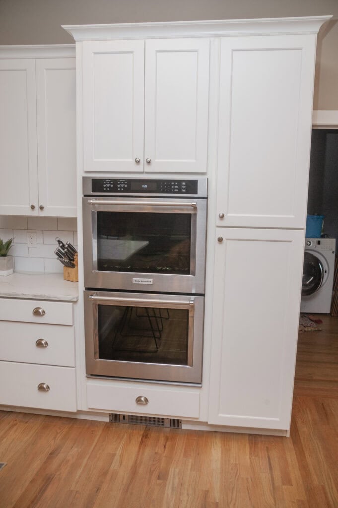 Double wall oven built into white kitchen cabinets, with silver handles, sits next to drawers and a countertop with a knife block. A laundry room with a washer is visible in the background. Hardwood flooring throughout.